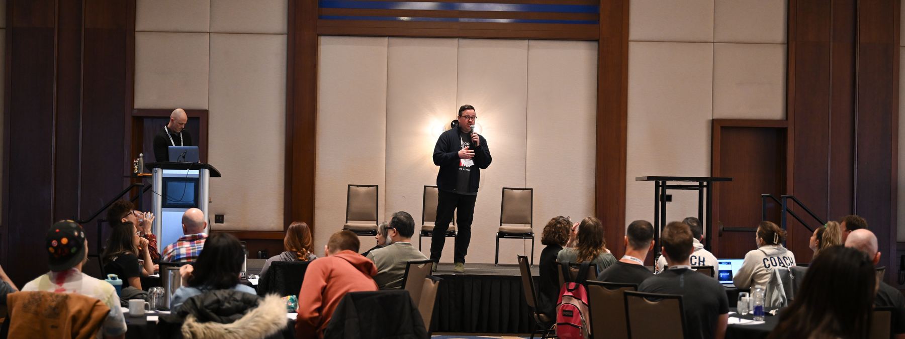 Michael on stage with a microphone, speaking to a seated ballroom audience.